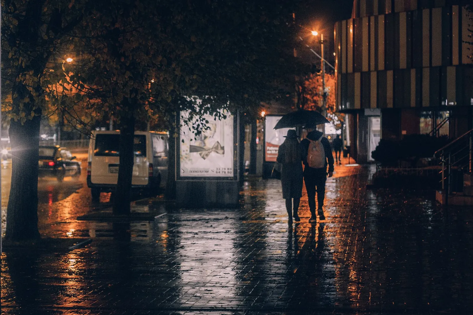 A couple walking together under an umbrella on a lamplit city street in the evening